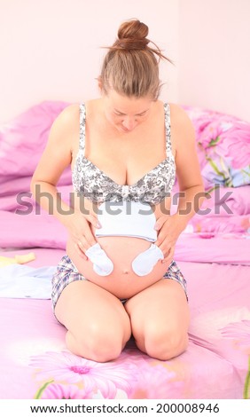 Pregnant woman tries on baby clothes to naked belly. Pregnant woman sitting on the bed with pink linen in the bedroom
