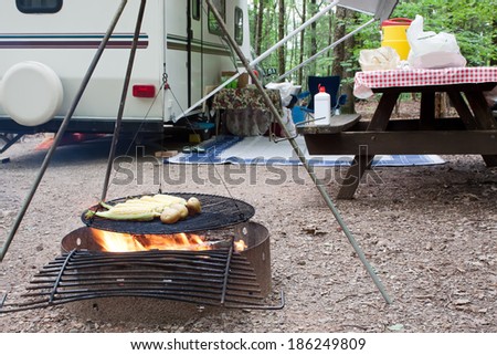 Corn and potatoes on an outdoor grill at a public park with a picnic table and camping trailer in the background.