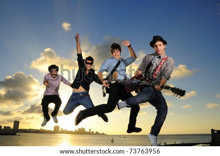 Band of young male musicians jumping with instruments against sunset sky background
