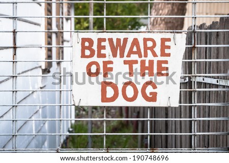 Hand painted red and cream sign wired onto a wire fence by the side of a house stating: BEWARE OF THE DOG