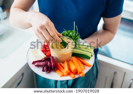 Top view close up man's hand dipping cucumber stick in hummus on the kitchen. Hummus served with raw vegetables on the plate. Healthy food lunch. Vegetarian and vegan food diet. Soft selective focus