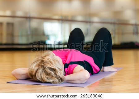 An mature blond woman in a bright pink yoga outfit rests peacefully on her back after exercising in a gym room.