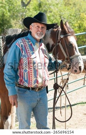 A older man in a black cowboy hat holds the bridle of a pinto horse.