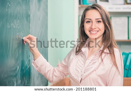 young tired female teacher holding open book above head at classroom blackboard background
