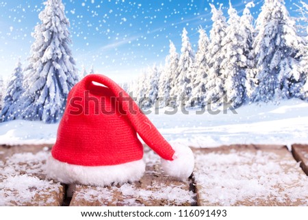red Santa Claus hat on a wooden table with snow outside with an amazing winter wonderland in blurred background, forest in winter