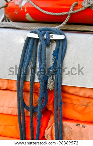 old blue ropes tied around a cleat on an old boat with orange life jackets underneath and a orange life buoy above it