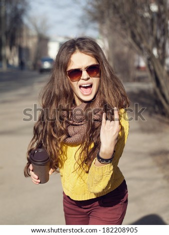 Beautiful brunette girl with takeaway drink walking down the street. Having fun, showing rock sign with her hand  and shouting (laughing). Urban city scene. Warm sunny weather. Outdoors