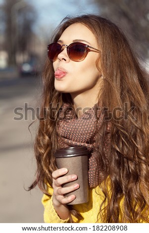 Closeup portrait of beautiful brunette girl with takeaway drink standing on the street. Having fun, showing tongue, looking happy. Urban city scene. Warm sunny weather. Outdoors