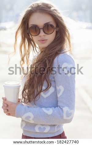 Portrait of a beautiful brunette girl in sunglasses standing on the street. Keeping drink in her hands. Warm sunny day. Outdoors