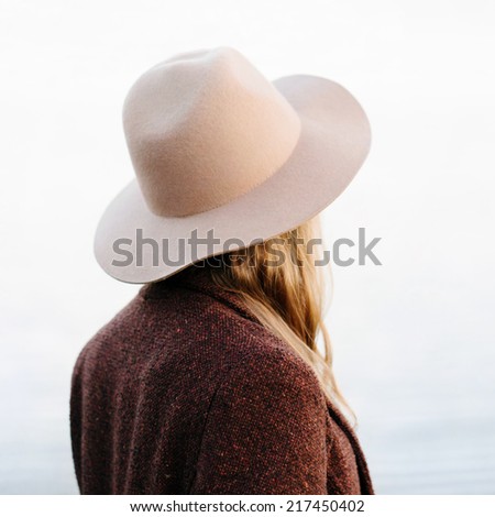 Portrait of blonde girl standing back to camera outside. Wearing hat and jacket. Outside