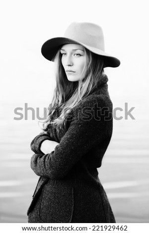 Portrait of a beautiful blonde girl standing on lakeside, wrapping up in jacket.  Wearing hat. Outside