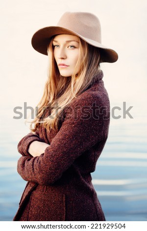 Portrait of a pretty blonde girl standing on lakeside, wrapping up in jacket.  Wearing hat. Outside