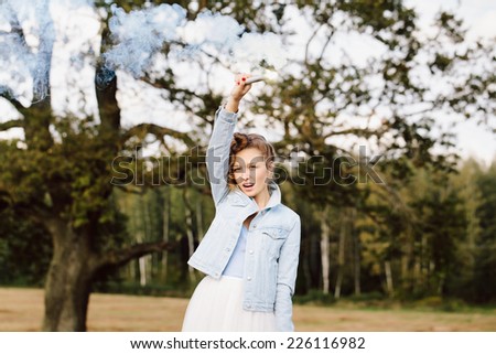 Portrait of beautiful blonde girl wearing casual clothes keeping flare with blue smoke in her hand. Nature background. Outdoors