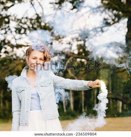Portrait of beautiful blonde girl wearing casual clothes and keeping blue smoke flare in her head. Nature background. Outdoors
