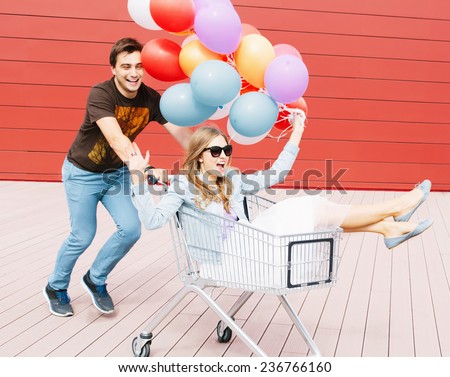 Portrait of two happy young people dating and having fun. Girl sitting in shopping cart and keeping color balloons in hand. Guy rolling trolley. Red background. Outdoors