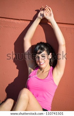 Young woman in sport wear rests on a wall and enjoys  listening music with white earphones. she is smiling and has raised arms.
