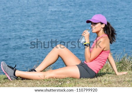 Beautiful brunette sportswoman resting and drinking water on grass with sea on background. Woman relaxing after exercising outdoors on summer sunny day.