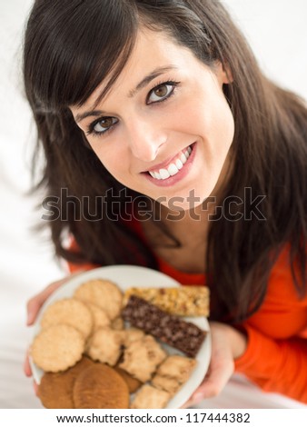 Cute brunette woman eating sweet cereal and chocolate biscuits in bed. Adorable and charming model, looking at camera and smiling.