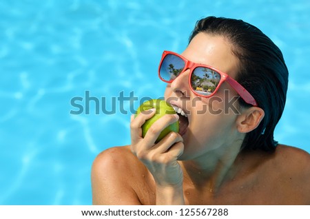 Young beautiful woman biting and eating an apple into swimming pool at hotel resort. Diet and summer concept with blue blur water background
