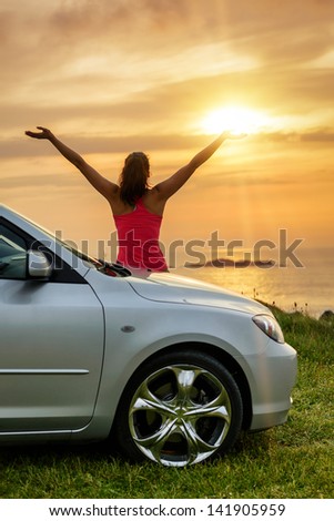 Free woman on car travel looking summer sunset sky and ocean. Female driver leaning on car bonnet raising arms to glow dusk sun. Traveling and freedom concept. Copy space.