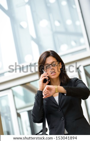 Stressed Business woman leaving corporate city building in a hurry looking time in her watch and talking on cellphone. Caucasian businesswoman working outside and being late for a business meeting.