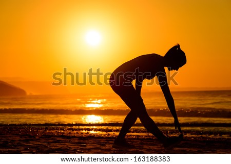Woman working out in beach at beautiful summer sunset. Fitness girl silhouette stretching legs on bright sun background.