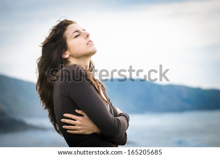Sad woman shivering and feeling the sea breeze. Nostalgic and emotional female hugging herself and feeling low on cold late summer or autumn day. Beautiful curly hair caucasian model.