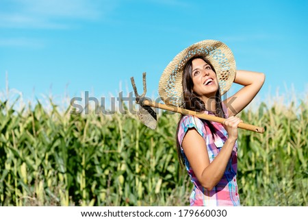 Successful female farmer carrying hoe in corn field and smiling. Countryside woman with work tool wearing straw hat and checkered shirt.