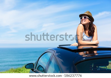 Relaxed happy woman on summer travel vacation to the coast  leaning out car sunroof with the sea on background.