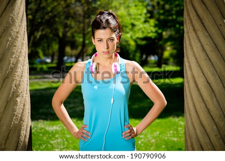 Strong fitness female athlete standing with determination and powerful attitude in city park. Woman on summer or spring workout outdoor.