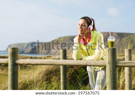 Pensive and relaxed fitness woman resting after exercising at beach on spring or summer. Fit brunette girl with detox smoothie drink. Playa de Xago, Asturias, Spain.