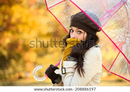Woman shivering in cold autumn and holding transparent umbrella. Brunette fashion girl covering her mouth with scarf outside.