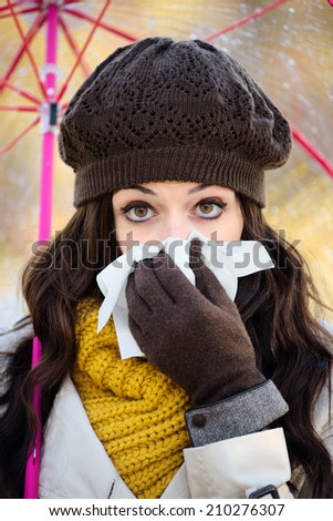 Woman with cold or flu blowing her nose with a tissue and sneezing under autumn rain. Brunette female wearing warm clothes, scarf, cap and gloves.