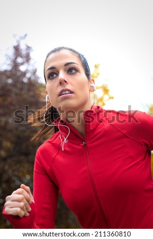 Female athlete on intense outdoor training in park. Beautiful serious runner exercising hard and wearing red sportswear and earphones.