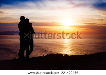 Young couple hugging and pointing to the sun on beautiful sunset or sunrise towards the tranquil sea. Love and romance concept.