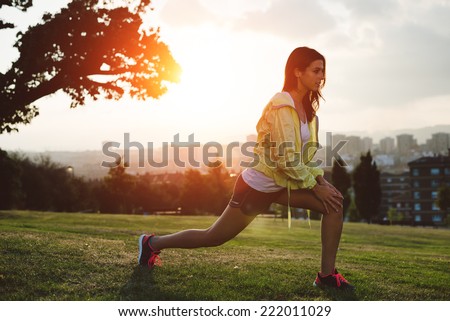 Female athlete stretching legs for warming up before running in city park on sunset or morning. Brunette  sporty woman exercising outdoor.