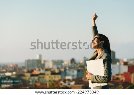 Successful young businesswoman raising arm celebrating business  or job achievement towards city background. Professional happy woman walking outside.