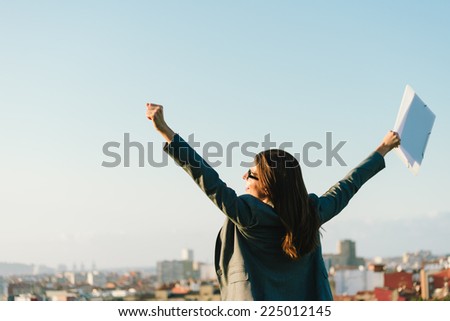 Successful young businesswoman with arms up celebrating business  or job success towards city skyline. Professional happy woman outside.