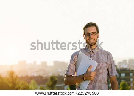 Successful professional casual man walking outside against city background. Happy smart looking person holding folder and smiling.