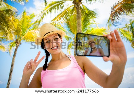 Woman on summer tropical caribbean travel taking selfie photo. Young happy brunette on beach vacation using her smartphone camera for self portrait with palm trees on background.