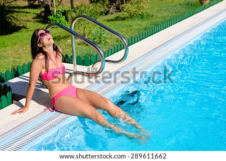 Beautiful woman in bikini sitting at poolside enjoying her summer  vacation.  Happy girl wearing sunglasses with feet under swimming pool water.