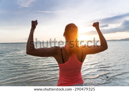 Sporty strong woman raising arms and enjoying freedom and success towards the sun and sea on sunset at the beach. Successful female athlete against the sun.
