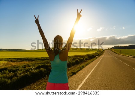 Woman celebrating running and training success on countryside road during sunset or sunrise. Female runner raising arms towards the sun.