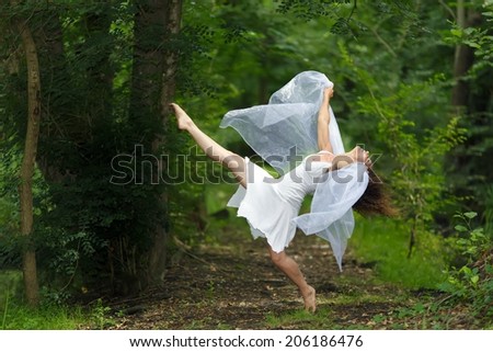 Mystical portrait of a beautiful graceful barefoot woman in a fresh white dress with her arms draped in filmy sheer fabric posing with one leg raised against a lush green woodland backdrop