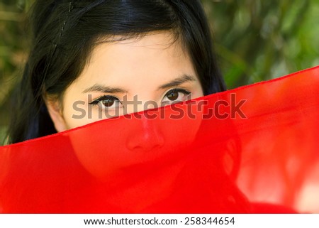 vietnamese woman peeking over red fabric at the camera so that just her eyes are visible, serious expression