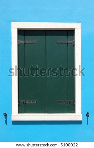 Window with closed green shutters, white outline, on a bright blue house in Burano Italy