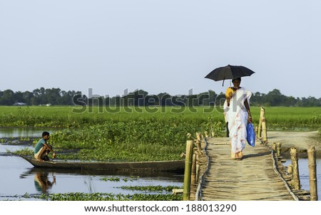 MAJULI, INDIA - AUGUST 27: Woman in sari and with umbrella crosses bamboo lattice bridge over lagoon as man in boat rows across and under the bridge on August 27, 2011, Majuli island, Assam, India.