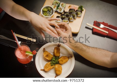 Proposal, Man giving ring to woman. Only hands and arms shown with sushi food at the table.