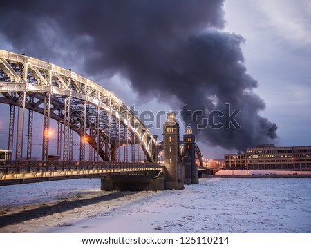 View on city fire - heavy black smoke polluting atmosphere and environment. Fire and smoke in the historical downtown over the river. Bolsheokhtinsky Bridge through the Neva River. St. Petersburg.