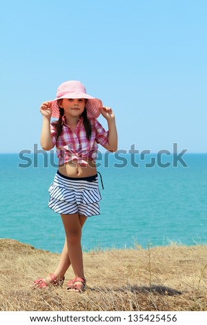 smiling girl in a hat and skirt stands with his legs crossed on a background of the sea